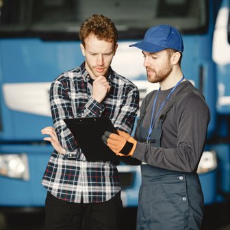 Ein Mann mit Handschuhen erklärt einem anderen, der nachdenklich schaut, Details auf einem Clipboard in einer Lagerhalle.